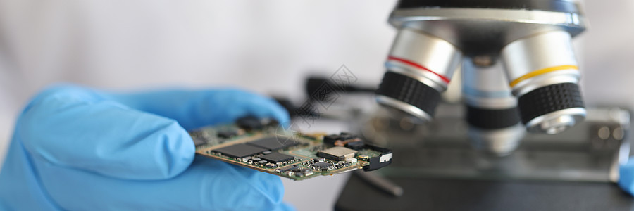手套工程师以显微镜为背景持有微型电路图片