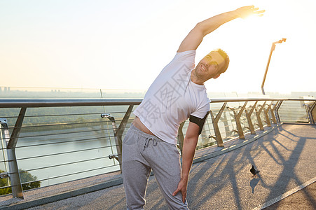中年肌肉健壮运动员在户外伸展手臂和肩膀 并做一个热身工作 在阳光明媚的夏季日子里享受早晨锻炼 站在一座城市桥上 欣赏美丽的大自然图片