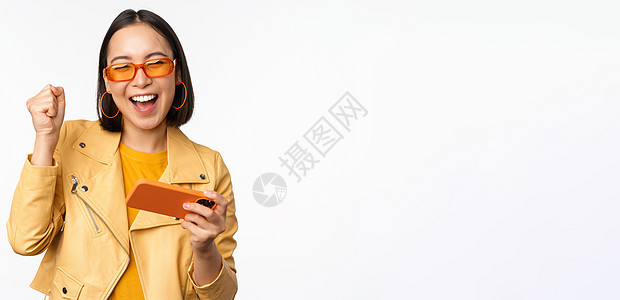时尚的韩国女孩用智能手机跳舞 欢笑快乐和微笑 站在白色背景之上的画面学生电话大学购物太阳镜技术商业工作室办公室工作图片