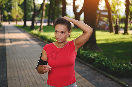 在早上沿着公园慢跑时 身穿红色 T 恤 手持智能手机的坚定女运动员站在小路上 系着马尾辫 有氧运动 积极健康的生活方式理念背景图片