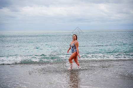 一位身穿泳衣的丰满女性在冲浪期间下水 独自在沙滩上 灰色的天空在云中 在冬天游泳女士姿势风暴身体泡沫海浪假期海岸海洋蓝色图片