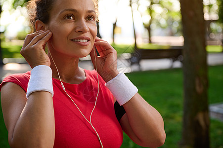 迷人的中年活跃的非洲裔美国女性 戴耳机 穿着鲜红色 T 恤和白色毛圈手镯的女运动员 准备进行户外锻炼和晨跑图片