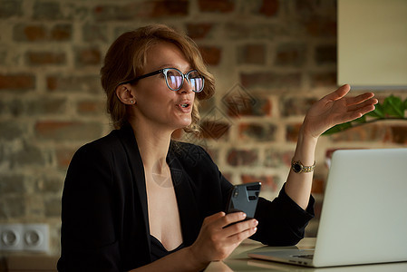 一个戴眼镜的年轻女子 远程工作 用智能手机在笔记本电脑上视频互联网商务网络手表长发女性电话女士会议图片