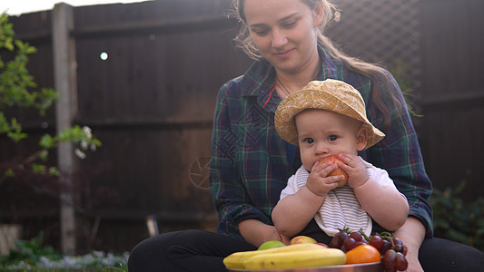 快乐年轻开朗的母亲抱着婴儿在绿草上吃水果 妈妈可爱的婴儿在后院花园里带着爱在户外玩耍 有父母的小孩 家庭 自然 生态概念女儿太阳图片