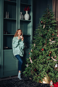 一个穿着自制衣服的漂亮女人 在家圣诞树附近喝着美味的茶或咖啡 很愉快的早晨饮料装饰女士女孩房间微笑享受风格假期房子图片