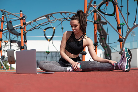 运动女运动员在夏季户外运动场用笔记本电脑健身健身房培训班练习灵活性运动鞋女孩下蹲瑜伽衣服冒充图片