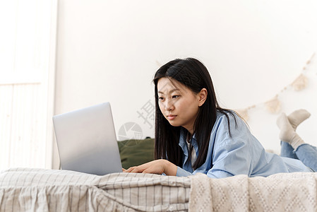 一位美丽的亚洲女性躺在床上 使用家里的笔记本电脑工作 一名年轻女子在家隔离工作 社交距离自我隔离微笑购物商业学生房间技术闲暇互联图片