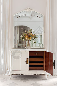 木制橱柜上的老式镜子和鲜花花束 带开门和抽屉的旧修复家具采用经典明亮的内饰 配有白色墙壁和窗帘图片