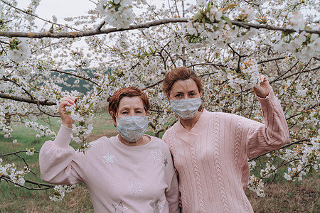 穿着粉红色毛衣医疗面具的妈妈和女儿正站在花朵樱桃花园里 在房子旁边有隔离概念图片