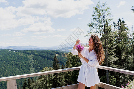 青山背景上花束着鲜花的迷人女孩 年轻女性穿着白色礼服 拿着粉红色花朵高山国家场景全景顶峰木头爬坡天空场地草地图片