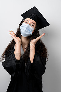 在冠状病毒 covid19 时期 戴着医用面具的单身女孩感到惊讶 硕士在毕业长袍和白色背景上的帽子 在线远程学习 在家学习 大学图片