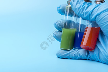一只戴手套的手在蓝色背景上 装着三个不同液体的小瓶子药店塞子技术员小瓶手指医生生物学微生物学生物玻璃图片