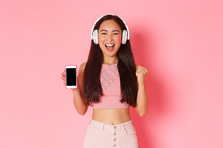 技术 通信和在线生活方式概念 华丽的亚洲舞女看起来乐观 戴着耳机听音乐时欢欣鼓舞 展示智能手机屏幕 粉红色背景图片