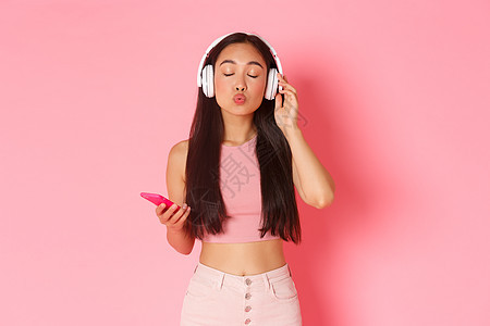 可爱而愚蠢的亚洲女孩的画像 穿着时髦的衣服 闭上眼睛 一边听着浪漫的歌曲一边噘嘴亲吻 戴着耳机享受音乐 拿着手机 粉红色的背景图片