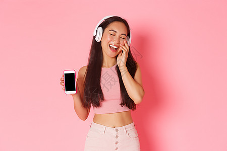 技术 通信和在线生活方式概念 华丽 无忧无虑的亚洲女性的肖像 闭上眼睛 微笑着可爱 一边戴着耳机听音乐 一边展示手机屏幕图片