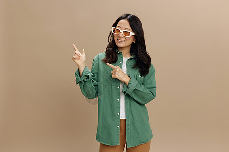 笑笑的韩国年轻女子 身着卡其青绿衬衫 时装眼罩和尖尖手指对准复制空间 在米色糊面工作室背景上被孤立 很酷的时尚提议 太阳镜广告概图片