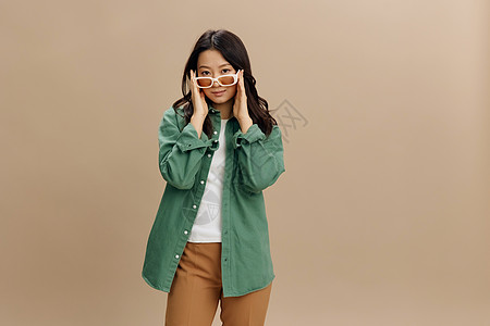 可爱的韩国微笑的年轻女子 穿着卡其绿色衬衫时髦的手戴眼罩 在米色面粉工作室背景上被孤立 很酷的时装报价 太阳镜广告概念美发护发素图片