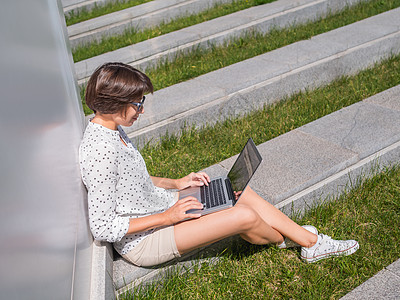女人坐在城市公园的长椅上 带着笔记本电脑 自由职业者在工作 学生在户外远程学习 现代生活方式 夏日风情 户外工作场所女性承包商网图片