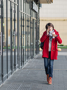 穿着红色大衣的女子由智能手机交谈 妇女走在街上拿起咖啡和纸支票的杯子图片