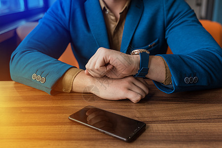 穿着蓝色夹克的商务人士看手表的时间 反面躺在手机或智能电话的木制桌子上的对面文字上图片