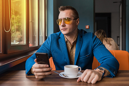 一位戴着墨镜的欧洲人的悲伤男人商务人士 一件蓝色夹克坐在咖啡馆的桌子旁喝咖啡休息时间 手里拿着智能手机 看着窗外图片