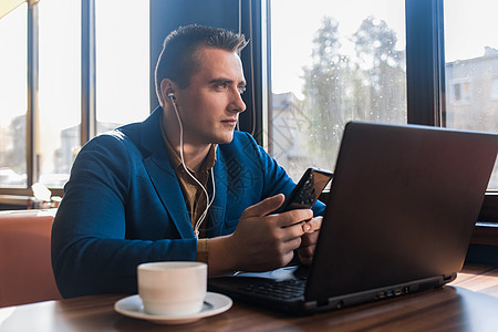 一位商务沉思的时尚商务人士 身着迷人的欧式西装 用笔记本电脑工作 戴着耳机听音乐 坐在窗边咖啡馆的桌子旁图片