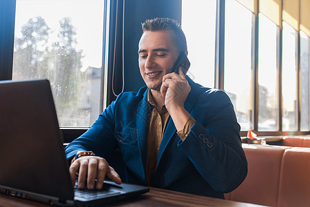 一位穿着迷人的欧式西装 时尚 积极微笑的商务人士在笔记本电脑上工作 用手机或手机交谈 坐在靠窗的咖啡馆的桌子旁技术办公室互联网商图片