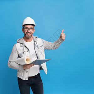 中年英俊建筑师 手持一卷墙纸和数字平板纸(c) 穿着时髦的散装衣 硬帽或头盔 在蓝背景上举起大拇指或完全孤立图片