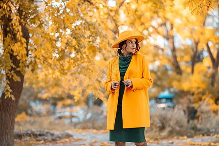 美丽的女人在秋天到户外散步 她身穿黄色外套 黄色帽子和绿色连衣裙 享受秋天天气的少妇 秋季内容微笑季节冒充落叶公园头发毛衣女士晴图片