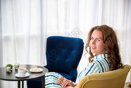 家里的年轻女人坐在窗前的现代椅子上 在客厅里放松 喝着咖啡或茶和糖果或沙漠 在新的工作日之前放松太阳巧克力房间房子商业女性杯子食图片