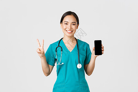 Covid19 医护人员和在线医学概念 微笑自信的亚洲女医生 身穿磨砂膏的医生展示和平标志和手机屏幕 推广咨询应用 白色背景看门图片