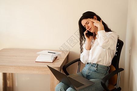 沮丧的女孩加班在膝上用笔记本电脑在线工作 并通过电话与同事交谈 疲惫不堪 压力过大 年轻漂亮的女人感到恶心和头痛生活桌子金融损失图片