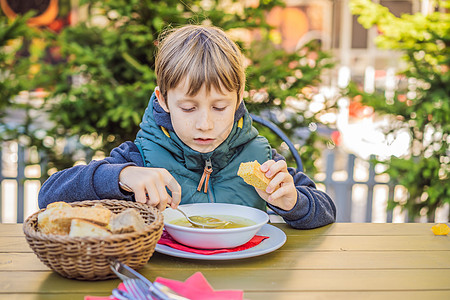 校园食堂可爱的小男孩在户外吃蔬菜汤 家庭厨房或学校食堂的金发小孩 可爱的孩子和健康食品 有机素食汤配土豆 番茄和蔬菜背景