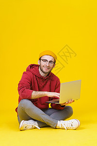 快乐的年轻时髦男子穿着红色连帽衫坐在地板上 拿着笔记本电脑学习或工作 黄色背景中带笔记本电脑的聪明自由职业白种人图片