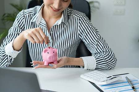 亚洲女商务人士的肖像是笑着在小猪银行里放一枚硬币 作为投资储蓄 财富和金融概念等图片