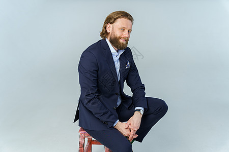 一个留着胡须和长发的英俊野蛮男人 一个坐在白色背景椅子上的时尚商务人士胡子选址蓝色戏服主义冒充裤子工作室情绪人士图片