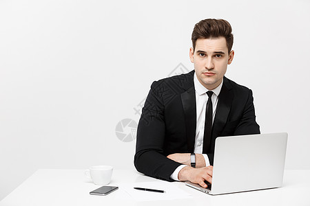 一位英俊的商务人士在他办公桌前的电脑上工作时手持智能手机的画像 他在办公室里 他的笔记本放在他面前 就像保险或银行经理一样互联网图片