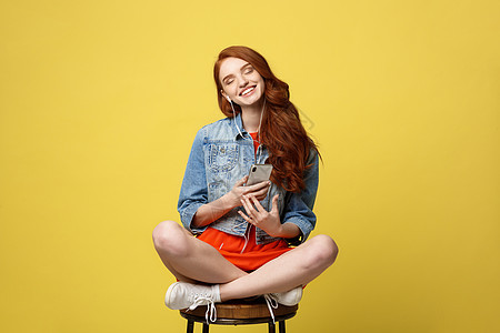 生活方式概念 长卷发红发的漂亮女孩在工作室用黄色背景生动地坐在木椅上 在电话机里听音乐 笑声微笑歌曲闲暇女士电话橙子学生椅子耳机图片