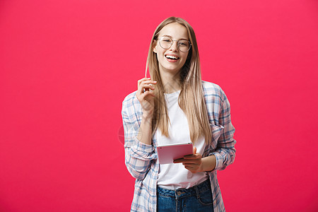 使用平板电脑 在粉红背景上隔离并观看相机的笑着女人的肖像屏幕白色学生快乐女性微笑软垫商业工作室技术图片