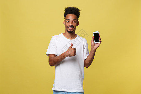 黄色背景中孤立的迷人年轻黑人肖像 手持空白智能手机 对着镜头微笑 竖起大拇指 感到快乐技术网络商务衬衫屏幕细胞手指人士男性拇指图片