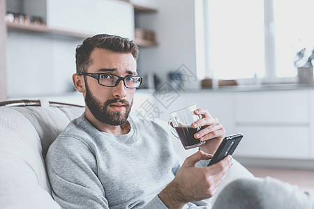 坐在舒服的椅子上坐着智能手机的青年男子 在互联网享受咖啡饮料杯子上网技术电话房子厨房图片
