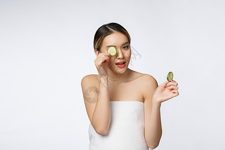 手拿着黄瓜片的年轻亚洲女人 在白色背景中孤立无援面具微笑治疗护理眼睛按摩相机黄瓜黑发皮肤图片