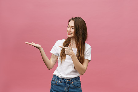 身着白衬衫的年轻女孩手对肩 用孤立的粉红背景展示产品手指微笑女孩女性粉色手势金发女郎黄色女士成人图片