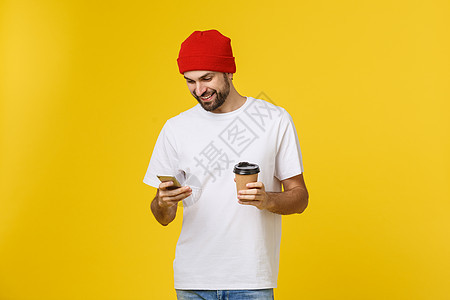 一个穿着休闲服的开朗年轻人的肖像站在黄色背景中 拿着手机 喝着外卖咖啡杯子快乐衬衫学生工具工作室男性人士技术自由职业者图片