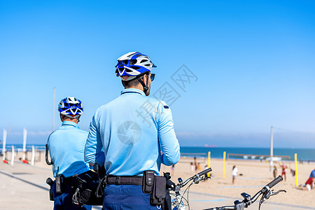 两名警察骑着自行车巡逻海边列车 人们在大西洋海岸的城市公共海滩晒太阳 美国海军陆战队民众热带男人长廊头盔车辆海岸天空街道运输图片