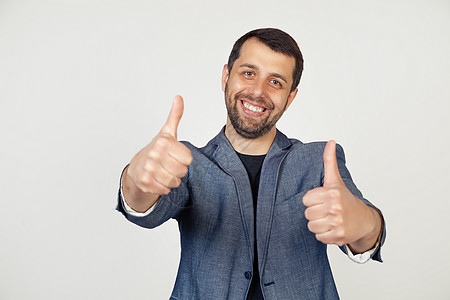 年轻的商务人士带着微笑 穿着夹克留着胡子 表示赞同 做出积极的手势 竖起大拇指微笑 为成功赢家的手势而高兴 灰色背景下的男人肖像图片