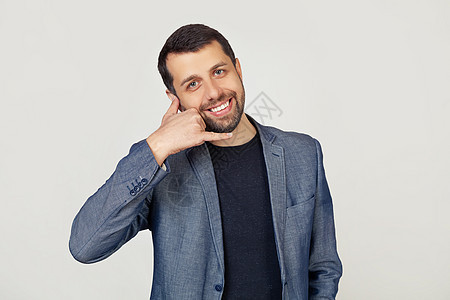 留着胡子的年轻商务人士穿着夹克 微笑着 用手和手指做着打电话的手势 就像在打电话一样 概念的迁移 灰色背景下的男人肖像图片