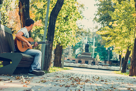 年轻人坐在公园的长椅上 用变调夹弹奏原声吉他 年轻有魅力的男人在最后一个阳光明媚的秋日假期享受现场音乐 使用复古镜头图片
