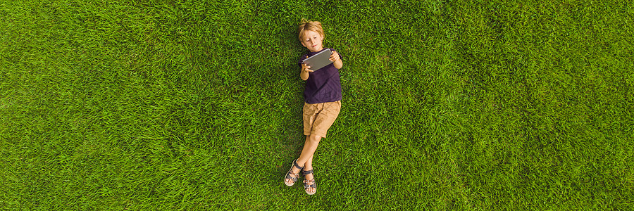 男孩躺在公园的草地上 看着平板电脑 照片来自无人驾驶飞机 四叉式BANNER Long ForMAT图片