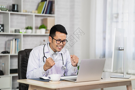 亚洲医生男人高兴地祝贺并使用笔记本电脑 男医生在家庭办公室医院工作 健康医院理念图片